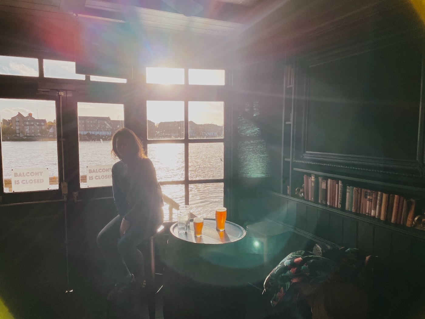 A girl sat at a table with beers in a pub, heavily backlit by a sunset