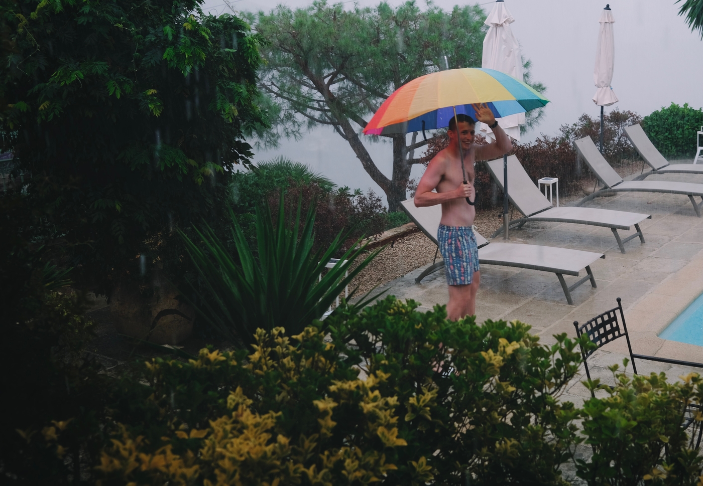 A man in swimming trunks in the pouring rain, holding an umbrella
